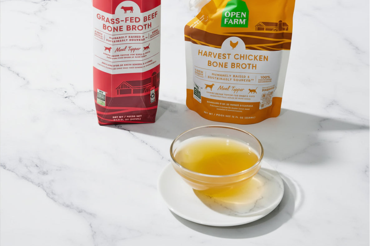 Two Open Farm bone broth pouches in red, orange, with a small glass bowl of golden broth in the foreground on a light marble surface.